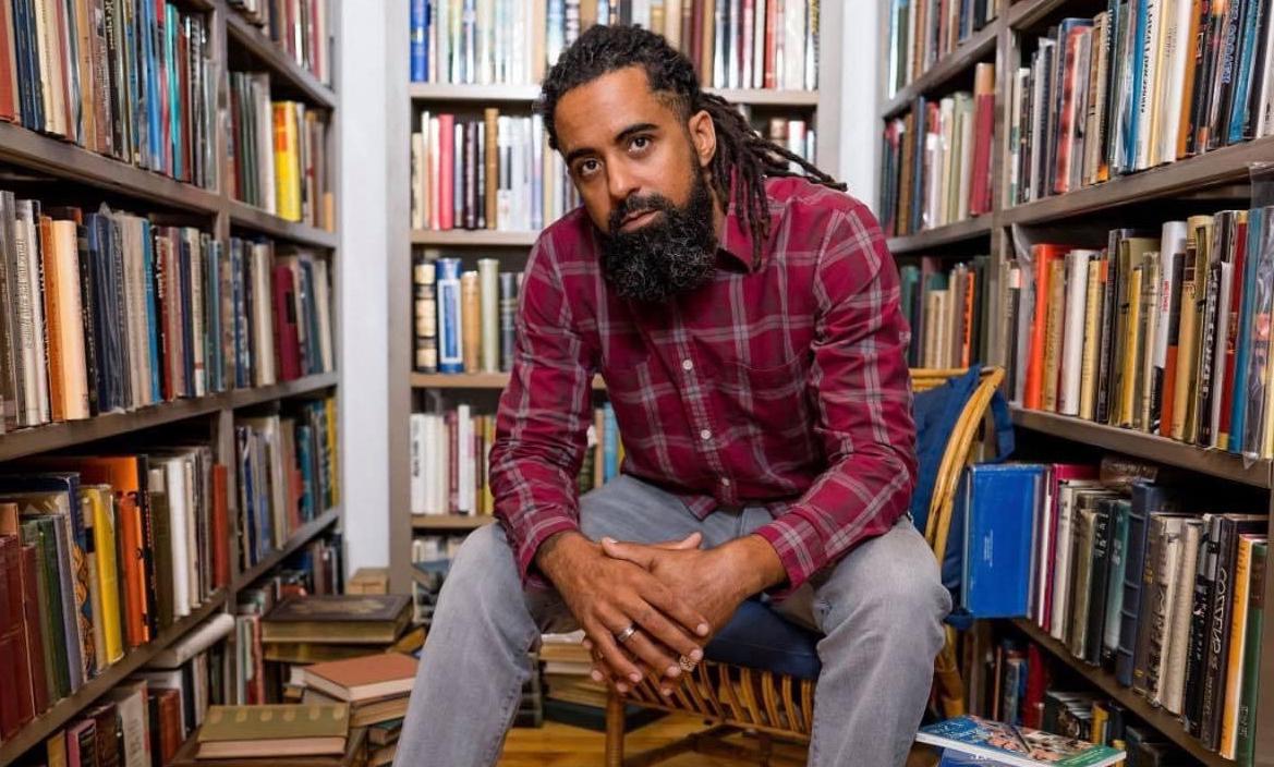 Jesse Kelley sits in front of shelves of books with books stacked on the floor